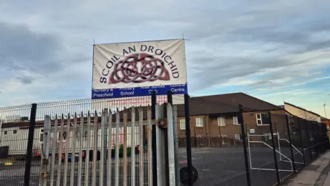 Scoil an Droichid sign with school behind it and grey sky. There's a playground with football nets behind the sign. 