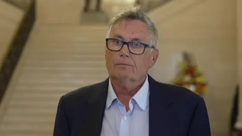 Robbie Marsland. He has short grey hair, wearing glasses, a navy blazer and white shirt. A marble staircase is behind him.