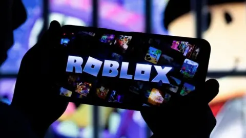 A pair of hands are holding up an iPhone which has the word Roblox in block capital letters on the screen. 