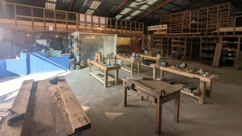 Ben McConnell A set of workbenches are in the middle of a warehouse. Hammers and woodchips can be seen on them. A large sign on the far side of the room reads "no smoking".