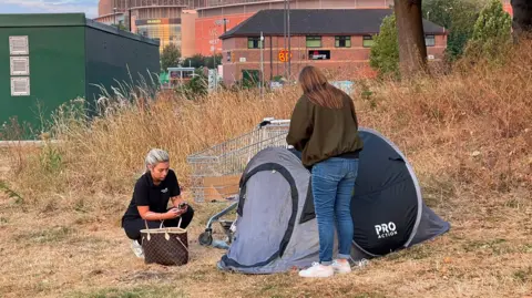 Two women stand beside a tent and a shopping trolley in a grassy area of a city centre. They are both typing on their phones. A shopping centre and office building can be seen in the distance.