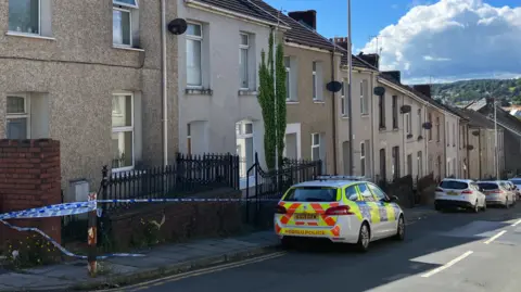 BBC Police car with other vehicles in Bigyn Street, Llanelli, with police tape along the pavement