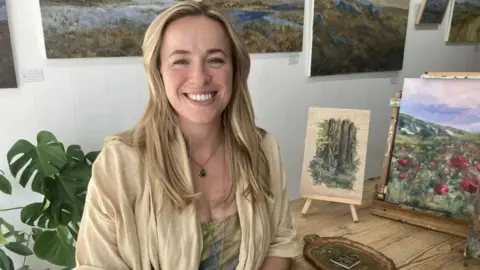 Lauren Taylor, with long blonde hair, beside paintings of trees and flowers.