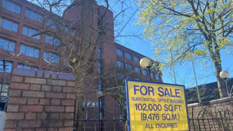 The entrance to a disused seven-storey office building. A black metal fence, attached to brick pillars, surrounds the site. Attached to the fence is a blue and yellow "for sale" sign. 