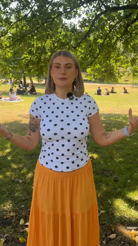 A woman with a blonde bob, white and black polka dot top and orange maxi skirt is standing with her arms outstretched.