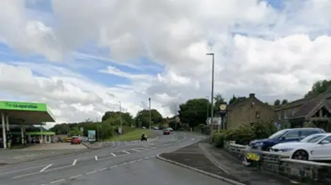 A junction featuring a petrol station opposite. Vehicles roadsigns and other street furniture can be seen. 