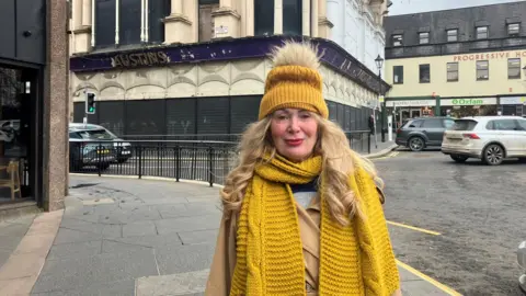 A woman in a yellow jacket and hat stands in front of a old derelict store in Derry city centre. A number of cars can be seen in the background. The woman has long blond hair and is smiling. 