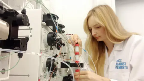 Race Against Dementia A blonde haired woman, wearing a labcoat, looks over some test tubes.