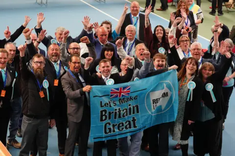 PA Media Two dozen Reform UK councillors celebrate on election night, with their hands in the air. They hold a sign saying Let's Make Britain Great Again.