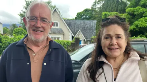 Pat Fitzgibbon and his wife Aisling Donohoe looking to the camera in Rostrevor. Pat has short, white hair and a beard and is wearing glasses, a navy jacket and a brown polo shirt. Aisling has long brown hair and is wearing a pink fleece and a gold necklace.