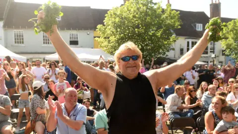 Alresford Watercress Festival Glenn Walsh in a black vest and sunglasses, his arms outstretched holding watercress in his hands as people cheer him on