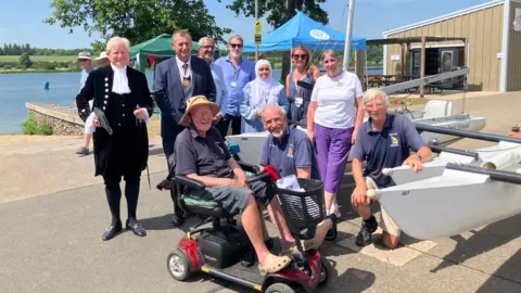 Shariqua Ahmed/BBC Group of people standing in the sunshine. On the far left is a dignitary in High Sheriff clothing, there is a gentleman in the front in a wheelchair