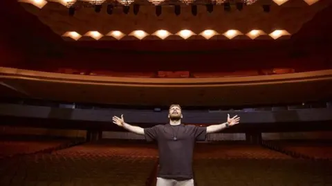 Anton Tymoshenko/Underground Standup Anton Tymoshenko stands with his arms outstretched in the middle of a red theater.