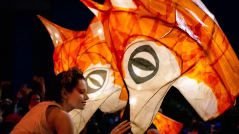 ANDREW MURRAY A woman leans down in front of a fox lantern.
