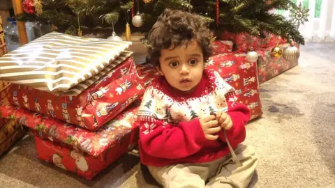Family handout A young boy sits in front of a mound of Christmas presents in front of a Christmas tree. He has curly brown hair and wears a red Christmas jumper with a tree pattern on it. He has cream trousers on.
