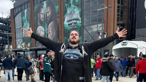 Jonathan Elgie A picture of Jonathan Elgie in a Wrestlemania top with his arms outstretched in front of the Wrestlemania sign