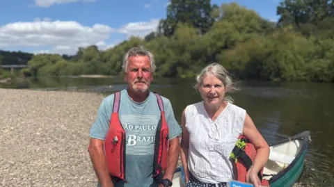 A man with grey hair wearing a light blue T-shirt is standing next to a woman with grey hair who is wearing a white top and standing by a canoe. A river and trees are in the background.