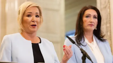 PA Media Michelle O'Neill who has blond hair tied back speaks into a microphone while Emma Little Pengelly who has long dark hair stands beside her.
