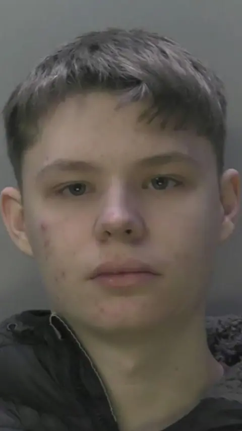 A mugshot of a boy with short brown hair and a black coat.