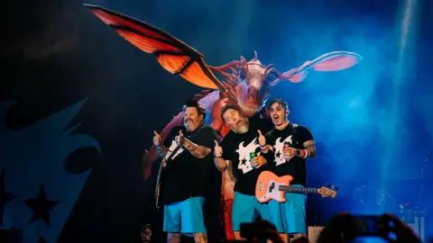Depot Live Frontman Jaret Reddick, bassist Rob Felicetti and drummer Gary Wiseman, from the band Bowling for Soup. The three band members are stood on stage whilst performing at Cardiff Castle. Behind them is a large red dragon, appearing over their heads. The band are all holding their thumbs up. Reddick and Felicetti are wearing guitars. All three members are wearing blue shorts and a black t-shirt with a white graphic print in the centre.