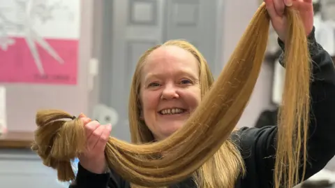 Louise Craig smiles as she holds her freshly cut hair tied with an elastic band.