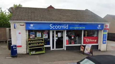 The front of the Scotmid shop has a a sign in blue. There are five bollards along the edge of the pavement in front of the shop.