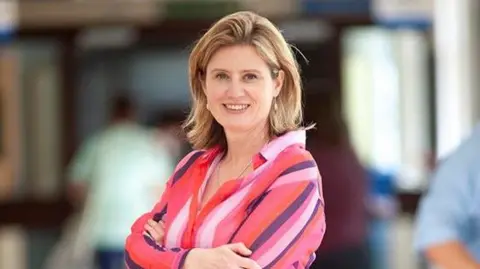 Norfolk and Waveney Integrated Care Board Dr Frankie Swords is standing to the side looking towards the camera. She is wearing a multicoloured striped shirt, and has arms crossed over her chest. She has shoulder length blonde hair and is smiling at the camera.