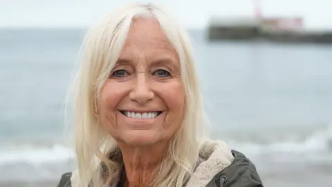 Susan George, a woman in her 70s with long blonde hair, stands on a beach, smiling at the camera.