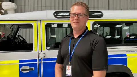 PC Linscott is standing in front of a police Land Rover Defender and is smiling. He has short blond hair, blue eyes and is wearing a pair of glasses. He is wearing a black police-branded short-sleeved T-shirt and a Devon and Cornwall Police branded lanyard which holds his identity card.