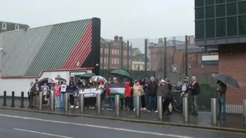 A group of about 30 pro-Palestinian protesters gathered outside Musgrave Street PSNI station in Belfast city centre.  Some are holding banners and umbrellas. A large Palestinian flag is being held up in the middle of the group.