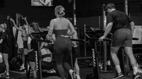 Kulture A black and white picture taken from behind as a large group of people exercise in the large warehouse gym, using weights and running and rowing machines. There is a long running mat on the floor which says 'Kulture' in capital letters, and metal shutters at the front of the building, which has tall ceilings and is very dark. 