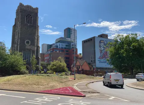 Andrew Woodger/BBC A grass verge off a roundabout is painted pink in the shape of a triangle with a white triangle in the middle of it. Beyond the roundabout are a church, modern high-rise buildings and an old industrial building with a mural on it. 