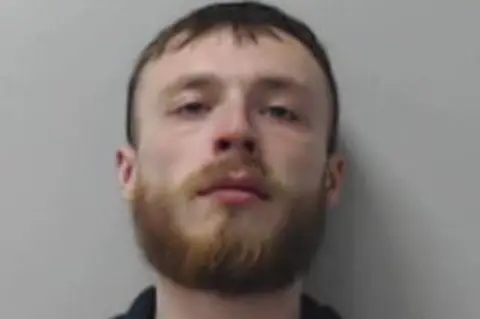 Police Scotland A head‑and‑shoulders portrait of a person with short hair and a full beard, wearing a dark jacket over a light top, photographed against a plain grey background.