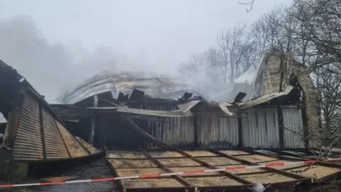 A partly collapsed blackened building with corrugated metal panels fallen or melted, and smoke emerging from the building - but no flames. There are deciduous trees to one side and a red and white emergency tape in the foreground. 