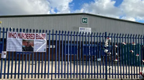 Handout A banner with the words 'Who Murdered Ellis?' tied to a blue fence in front of a warehouse. There are also bunches of flowers tied to the fence