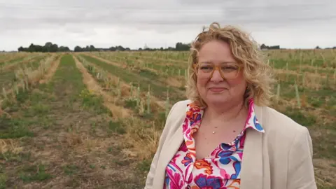 O femeie pe un câmp, lângă copaci plantați. Poartă o jachetă crem, o cămașă cu motive florale și un colier în jurul gâtului. Are părul scurt și creț și poartă ochelari.