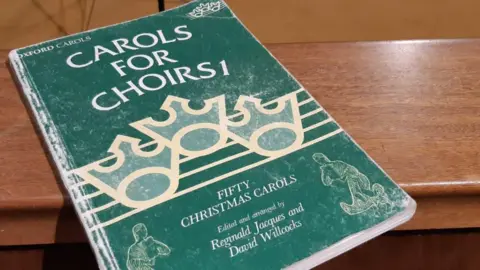 A green book reading "Carols for Choirs 1" - FIFTY CHRISTMAS CAROLS - Edited and arranged by Reginald Jacques and David Willcocks. It is places on a wooden surface.