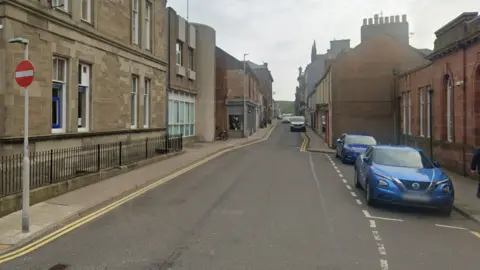 Google A general view of Commerce Street in Arbroath. There is a stop sign and two parked blue cars. There are several buildings on either side of the road. A van is parked on the right hand side in the distance.