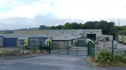 A grey factory with a collection of green and black lorry's and vans sitting in the forecourt, it is sorrounded by green metal fencing