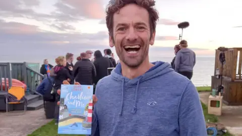 Mark Lowery A man holds a book in one hand. He is smiling widely. He has thick, short curly hair and wears a blue hoodie. A film crew can be seen in a group behind him talking. They are on the coastline.