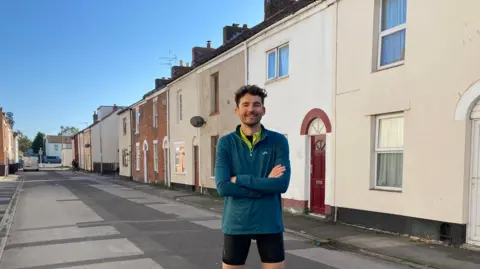 BBC Charlie Taylor is standing on a road with a row of terraced houses behind him. He is wearing cycling gear and a blue fleece. He has crossed his arms and is smiling.