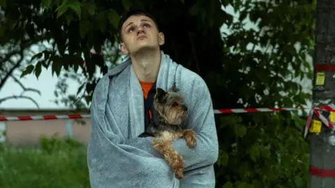Reuters A resident holds his dog as he looks an apartment building that was damaged during a Russian drone strike in the Ukrainian capital Kyiv