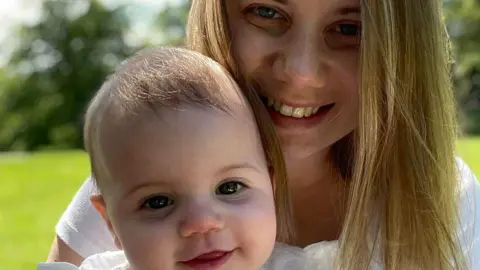 Chris Tuczemskyi Rebecca Ableman holding her daughter Autumn on a sunny day. Rebecca has long blonde hair and is smiling into the camera. Autumn is a baby in the photograph and is also staring into the camera. 