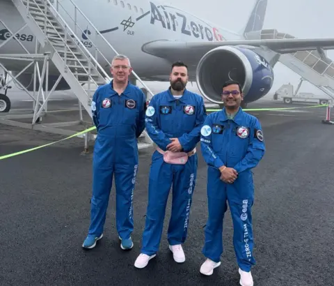 University of Glasgow 3D printing team Colin McInnes, Gilles Bailet, Satyam Bhatti, wearing blue flight suits marked with the European Space Agency Zero-G Team logo in white, stand on the tarmac in front of the Air Zero G plane.