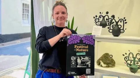 Lois Gunn wearing round dangly earrings, necklace, black jumper and blue trousers with a brown belt, holding up a Festival of Nature poster. She is looking at the camera. Behind her is some shelving on which there are ceramics. 