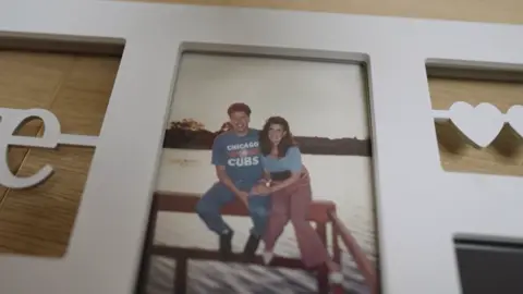 A photograph of Geraldine and her husband Glenn sat on a decking area overlooking a large lake. She has black curly hair and is wearing a blue top and pink trousers. Glenn is wearing jeans and a light blue t-shirt.