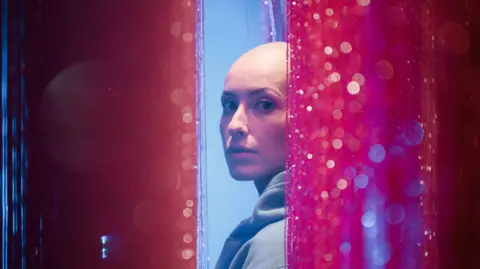 Emily Glaze is turning to look at the camera side-on from between pink glittery curtains. She has a bald head and is wearing a light blue hoodie. 