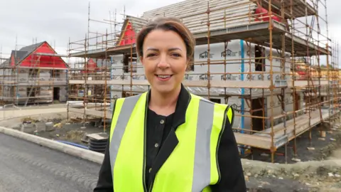 Liz Kimmins stands in front of a house currently under sconstruction. It is covered by scaffolding. Kimmins is wearing a high viz yellow jacket and black blouse. She is smiling and has short brown hair