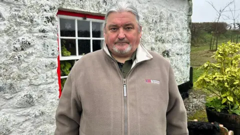 A man with a grey ponytail and goatee stands outside an old white cottage, with bright red framing on the window.
He is wearing a light brown fleece, with a red helicopter badge on the collar
Beside him is an old wheelbarrow carrying turf.
