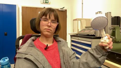 Kate is pictured seated in a laboratory setting. She is holding up a shower head wrapped in white material with workshop equipment, measuring devices and storage cabinets visible in the background.